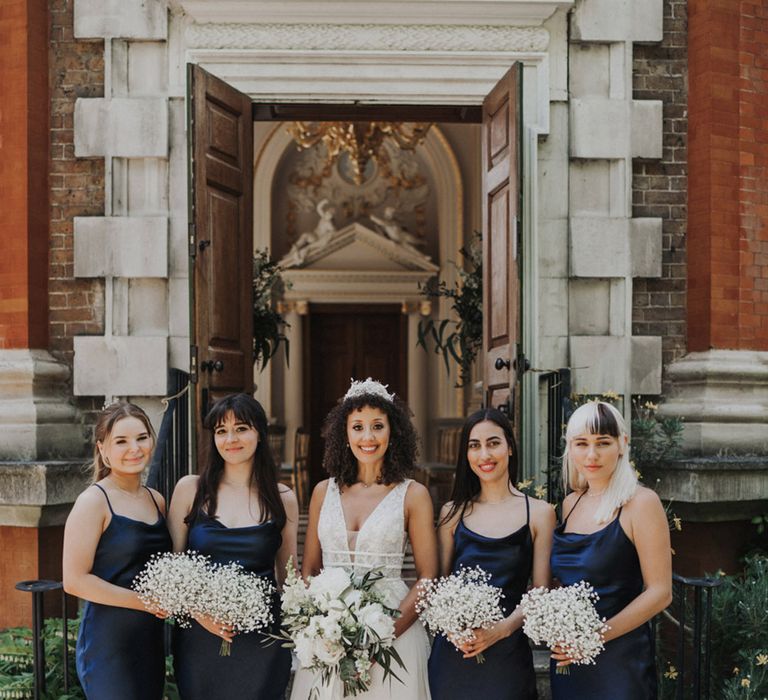 Bride wears bridal crown in her naturally curly hair and stands beside her bridesmaids in navy midi bridesmaid dresses 