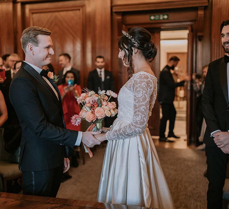 Groom in classic black tux with bowtie and rose boutonniere holding hands with bride in  taffeta material princess wedding dress with long lace sleeves and gold, pearl and emerald bridal crown holding bouquet with light pink poppies, orange garden roses, eucalyptus, Christmas red berry garlands, dried flowers and sage green and orange ribbon