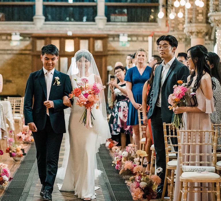 Colourful florals line the aisle as bride in drop ruffled veil holds colourful bridal bouquet as she walks down the aisle 