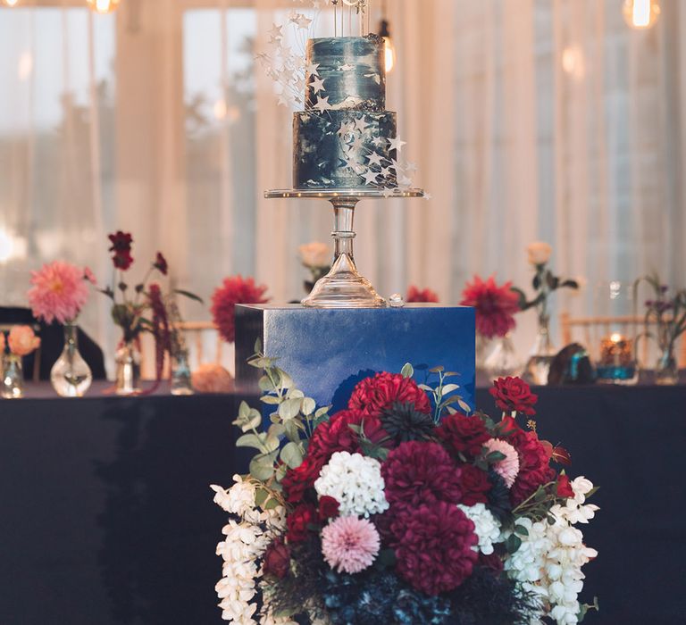 Two tier midnight blue wedding cake with white stars and moon cake toppers on a glass cake stand with red and white wedding flowers 