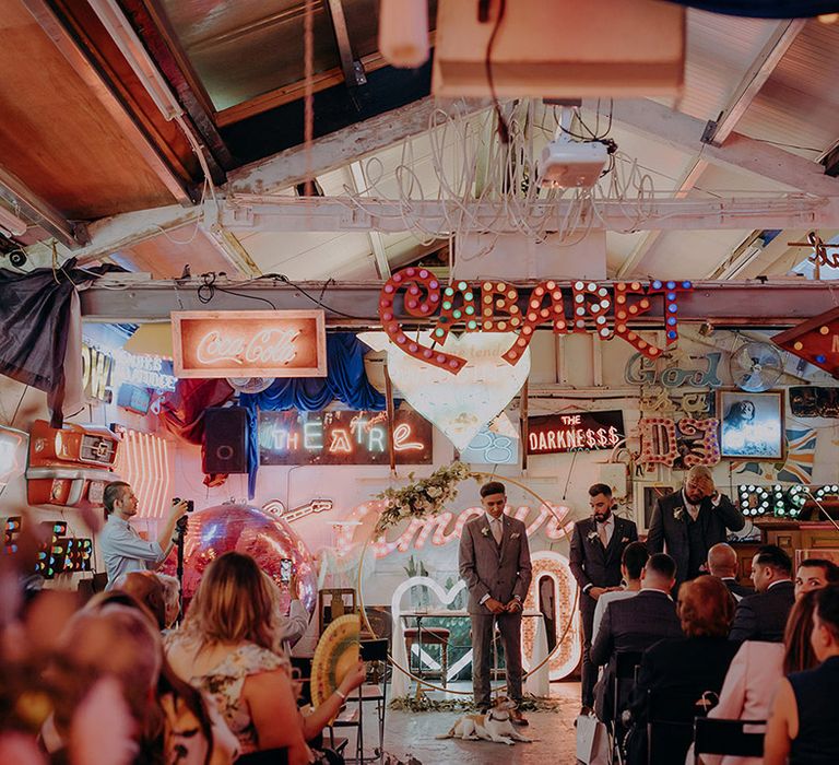 Groom in grey three piece wedding suit with yellow suit and yellow pocket square waiting at the end of the alter with numerous bright neon signs, LED signs and large wooden circular arch with rose decorations attached 