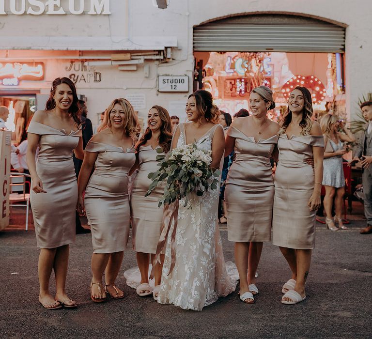 Bridesmaids in midi length off-the-shoulder satin silver bridesmaid dresses holding white rose floral hoop and bride in sleeveless plunge lace wedding dress holding white rose, eucalyptus and foliage bouquet 