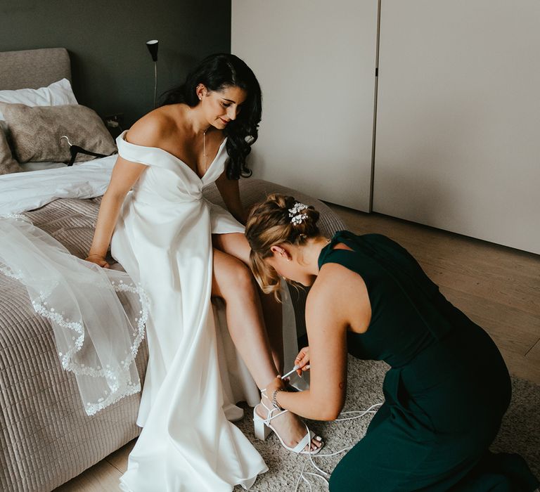 Bridesmaid in green bridesmaid jumpsuit helps bride with her strappy white bridal shoes with block heel