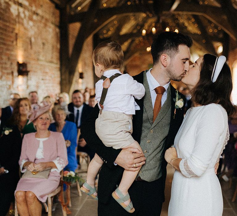 Groom in grey tweed waistcoat and orange tie holding his one year old toddler kisses the bride who wear a high neck wedding dress with a pearl headband and thick white headband