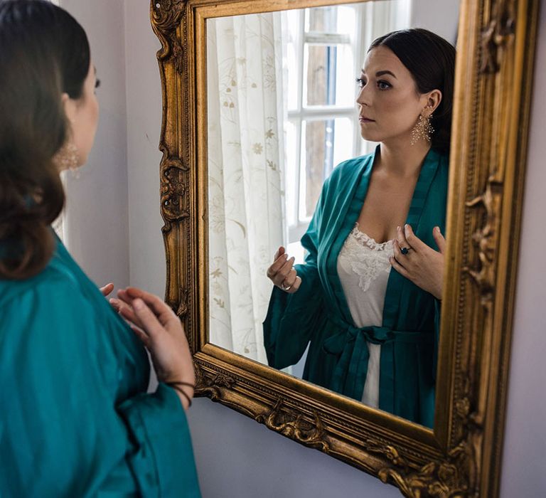 Bride in a turquoise satin robe and white pyjamas looking in the mirror as she gets ready for the wedding 