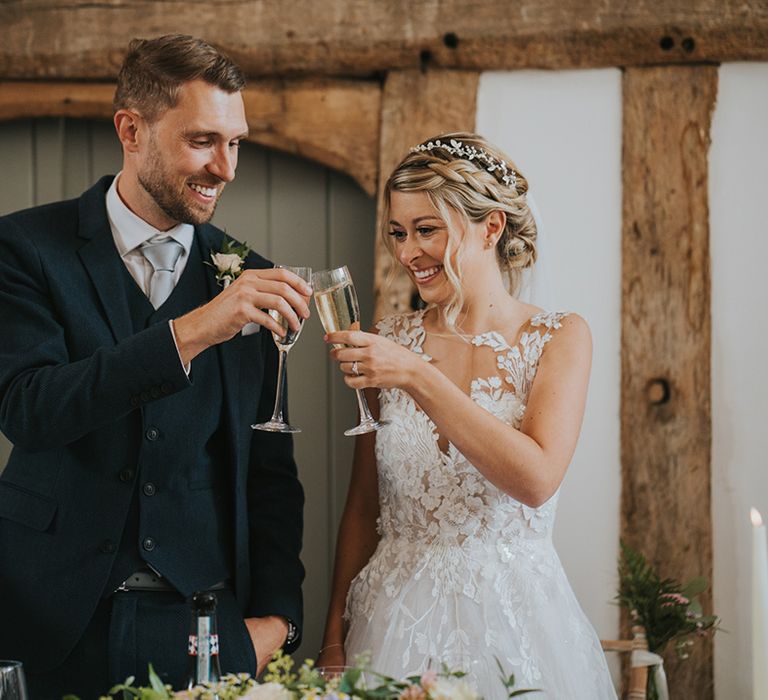 Bride with blonde braided hairstyle with lace wedding dress with groom in navy suit cheers their glasses of champagne together 