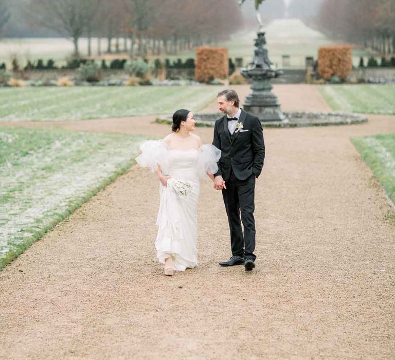 Bride in strapless wedding dress with puff tulle sleeves and groom in classic black tuxedo, black bowtie and boutonniere walking the grounds of St Giles House