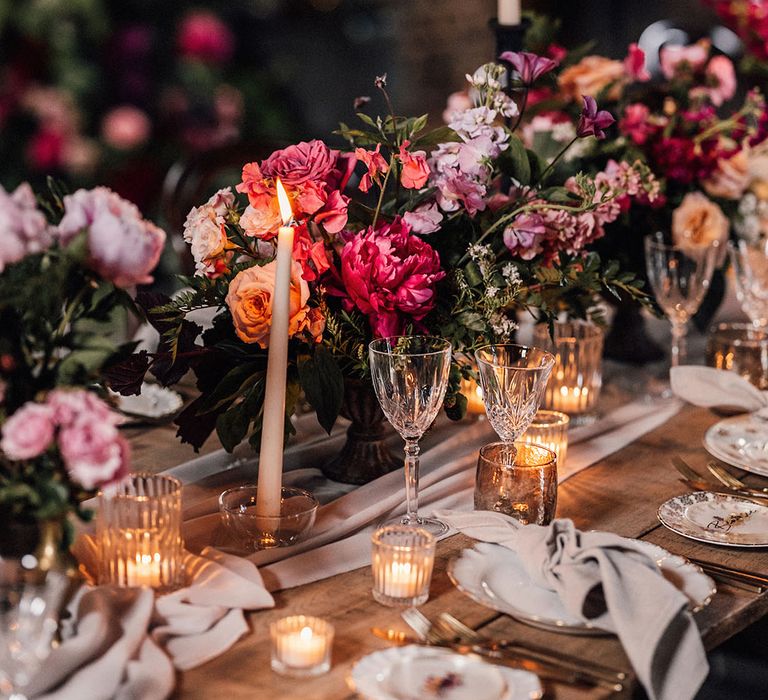 Pink peony and rose floral arrangements on the table with draped table runner, white taper candles, and cosy tea lights, with crystal glassware and light grey napkins 