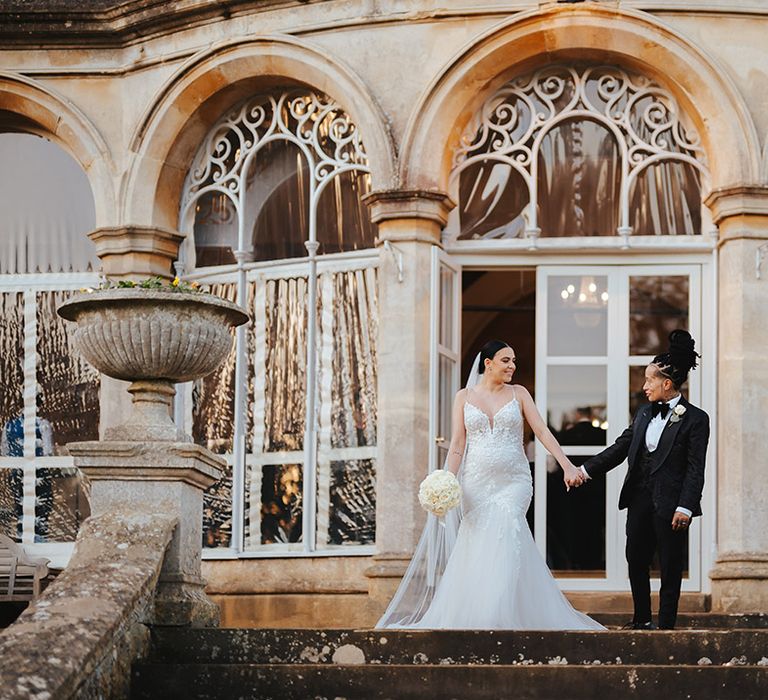 Bride in black snakeskin print tuxedo with black bowtie and white rose boutonniere holding hands with bride in lace sleeveless v neck Enzoani wedding dress with tulle finish holding white garden rose bouquet outside Grittleton House wedding venue 