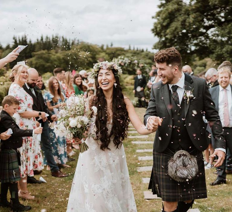 Groom doing confetti exit at wedding at The Barn at Harburn wearing dark grey blazer and waistcoat, dark tartan tie with white flower boutonniere, dark grey and blue tartan kilt and wedding sporran with bride in celestial 3d appliqué sheer overlay wedding dress and bridal flower crown holding large white rose, peony, dried flower and foliage bridal bouquet 