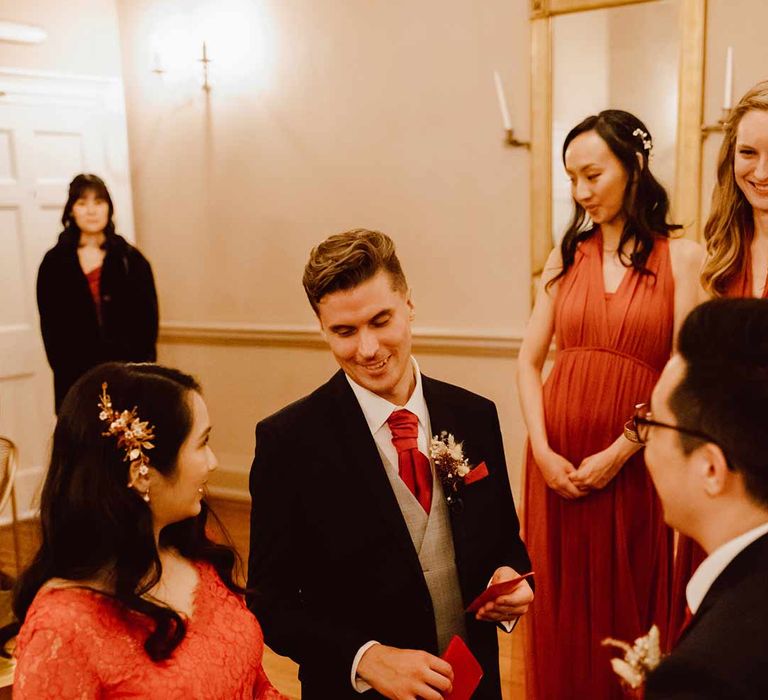 Bride in red lace wedding dress with long sleeves, red and gold hair accessories and golden closed toe wedding heels smiling at groom in deep blue grooms blazer, grey waistcoat, red tie, red pocket square and mixed dried flower boutonniere at The Orangery Maidstone wedding