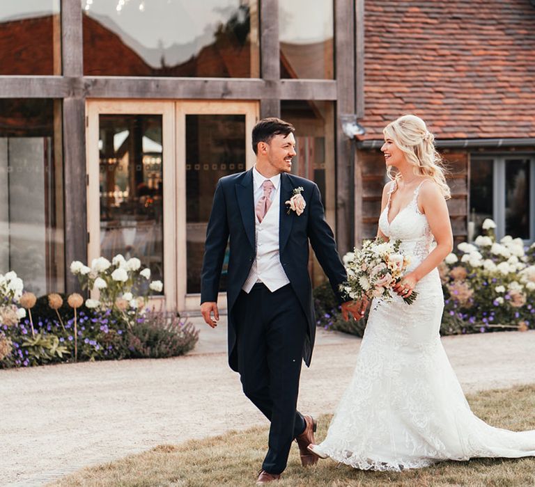 Groom in navy morning suit with light grey waistcoat and pink tie walking along with the bride in a lace wedding dress at rustic barn wedding venue 