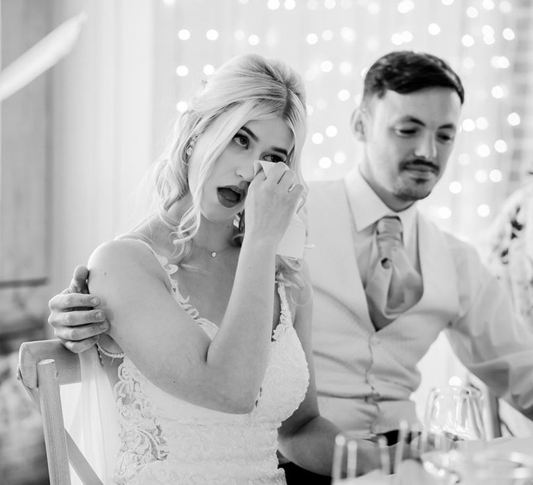 The bride wipes her eyes as she gets emotional listening to the wedding speeches with the groom putting his arm around the bride 