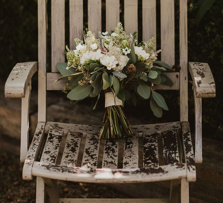 A white wooden chair holding the bridal bouquet with white flowers and foliage 