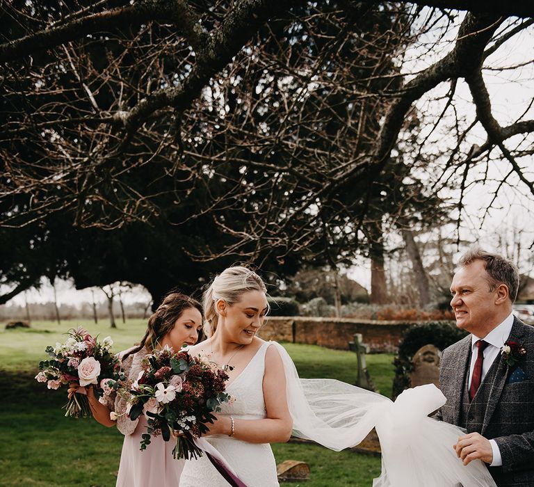 The bridesmaids and father of the bride help the bride with her wedding dress and veil as they walk to the church