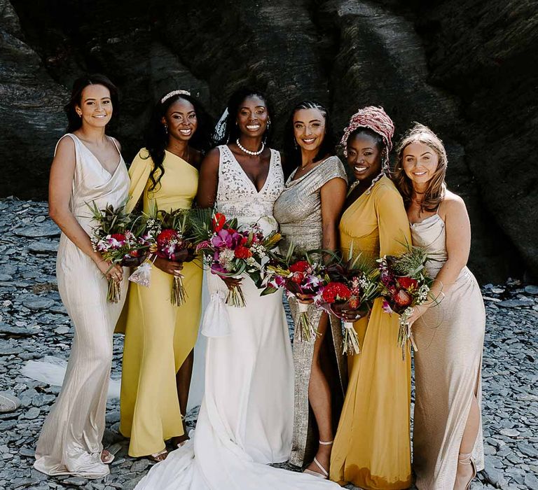 Bridesmaids in yellow, silver and gold mismatched bridesmaid dresses standing with bride in v neck sleeveless lace Pronovias wedding dress, cathedral length veil and silver sparkly bridal jewellery on Tunnels Beaches holding colourful tropical African bridal bouquets