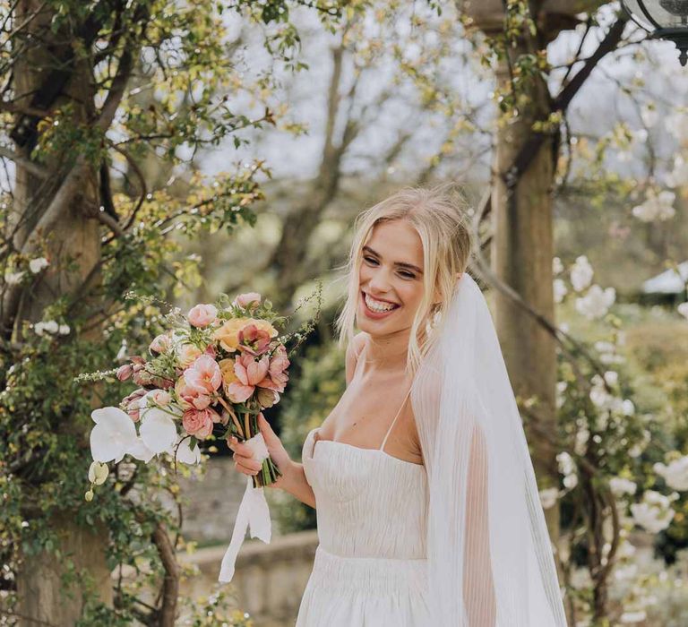 Bride holding blush floral bridal bouquet wearing Watters strappy sleeveless princess wedding dress and chapel length wedding veil 