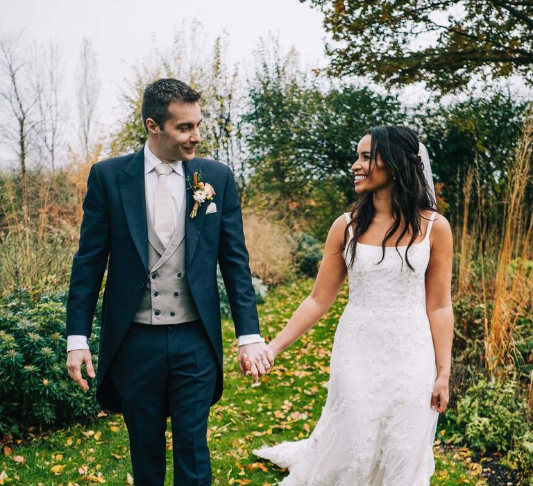 Bride in a-line tulle wedding dress with beaded lace applique with petals and a square neckline with side tulle cutouts and church length veil holding hands with groom in navy grooms morning suit with grey waistcoat, off-white tie and mixed wildflower boutonniere at South Farm wedding