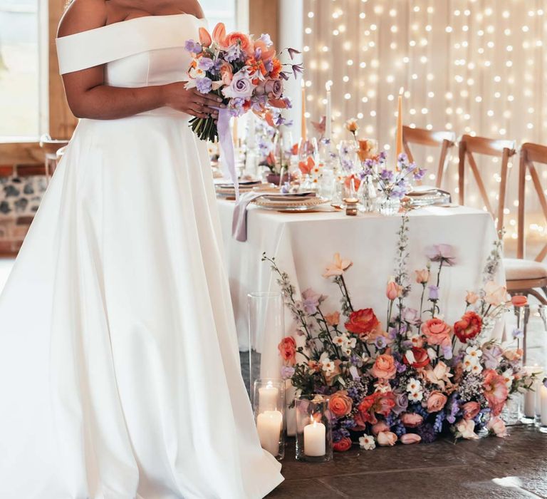 Bride in off the shoulder wedding dress with puddle train holding orange, lilac and peach mixed flower bridal bouquet standing by peach and lilac wedding tablescape with spring flower centrepieces, lilac napkins, tinted glassware and gold cutlery and crockery on white tablecloth with flower arrangement with peach garden roses, white peonies, lilac sweet peas, bluebells and wildflowers at Rackleys Barn