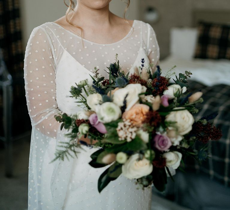 Bride in sheer long sleeve dotted wedding dress carrying white, purples and red wedding bouquet of wildflowers 