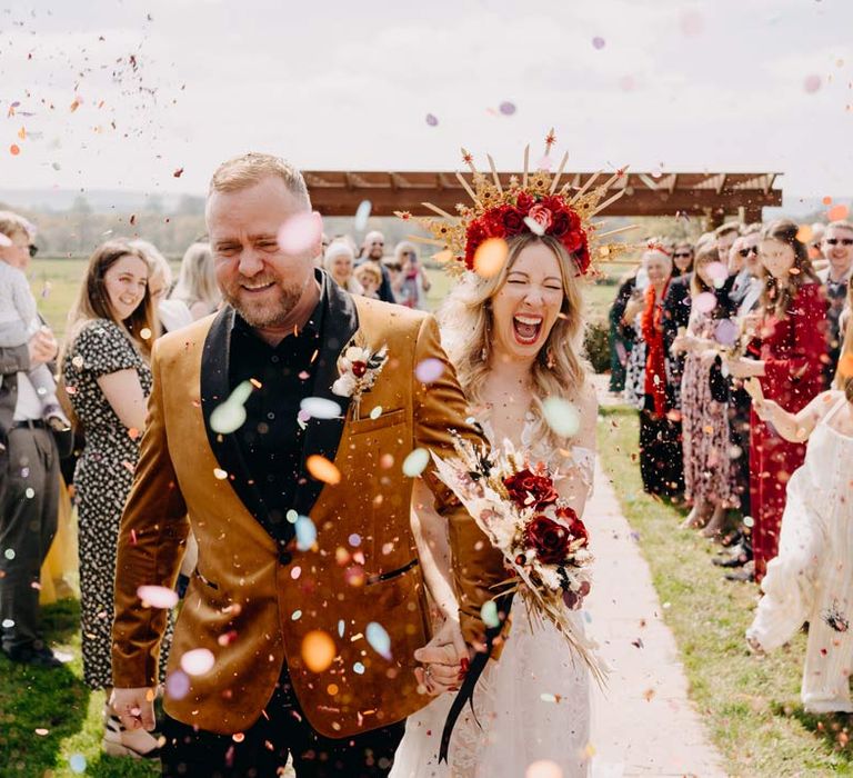 Bride in off shoulder lace wedding dress with corset style top and red rose and gold halo bridal crown holding red rose, pampas grass and dried flower bridal bouquet doing colourful confetti exit with groom in crushed velvet burnt orange grooms blazer and dried flower boutonniere at Southlands Barn