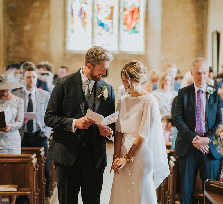 Groom in three piece wedding suit with green tie looking through the order of service book with the bride in a classic wedding dress with overlay for church ceremony 