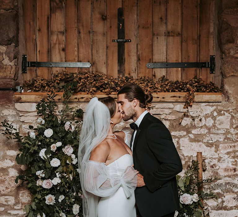 White wedding flower column arrangements frame the altar behind the bride and groom as they share their first kiss as a married couple 