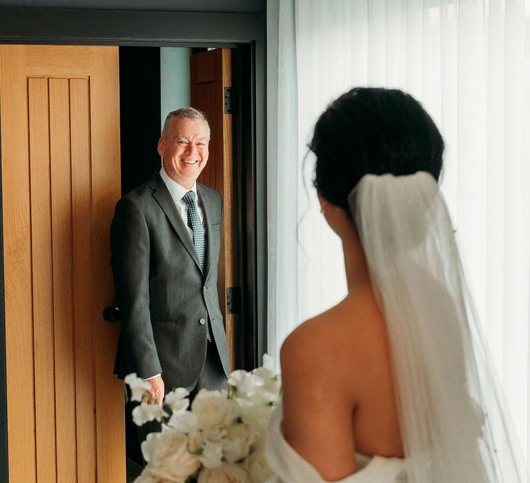 Father in charcoal wedding suit smiles at bride in first look photo