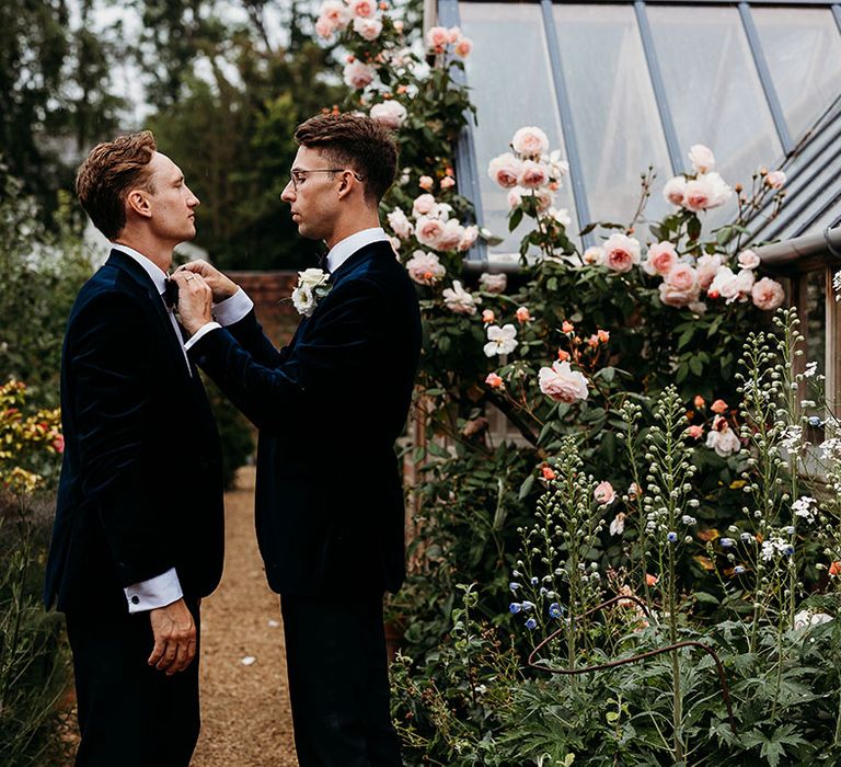 Groom in blue tuxedo helps the other groom with his bow tie 