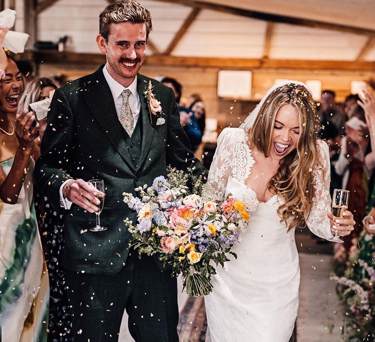 Confetti exit for the bride long sleeve traditional lace gown and groom in a dark green suit 