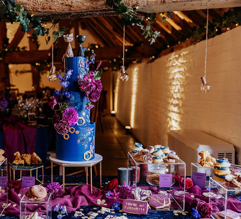 Dark blue wedding cake decorated with cat cake topper on a dessert table with mini ice cream cones, biscuits, flapjacks and more 