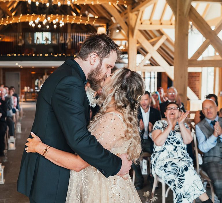 The bride in a gold wedding dress shares her first kiss as a married couple with the groom in an all black wedding suit 