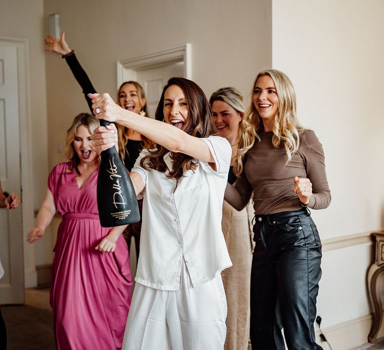 The bride in white satin pyjamas pops a bottle of champagne as they bridal party start getting ready 