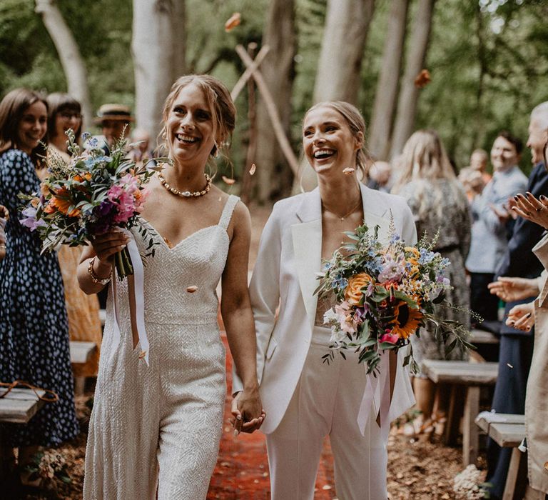 Bride in suit and bride in jumpsuit walk back down the aisle together as a married couple 