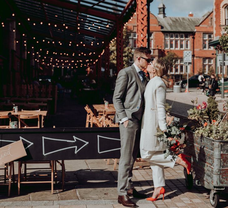 Bride in Massimo Dutti jumpsuit and Ivory coat and groom in grey check suit kissing 