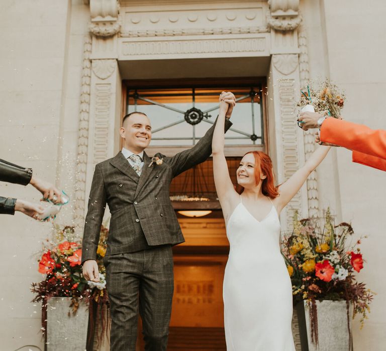 Confetti moment for bride and groom outside city wedding. Holding up their arms in celebration. Bride wears a simple silk wedding dress groom wears check suit. 