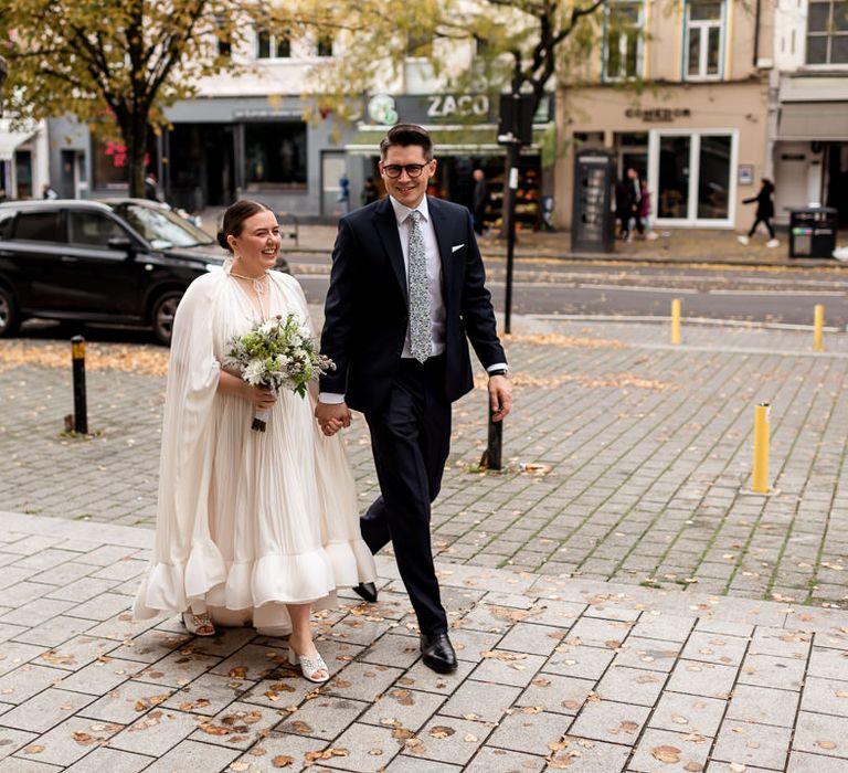 Groom in a navy suit holding hands with his bride in a Lanvin wedding dress, bridal cape and mules 