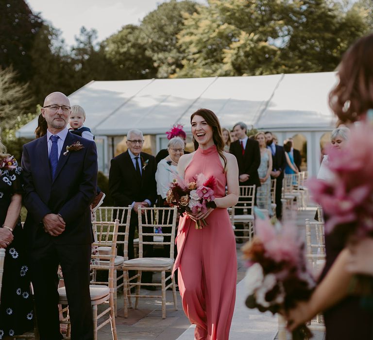 Bridesmaid in pink Rewritten bridesmaids dress holding pink and white rose and pampas grass bouquet walks up the aisle