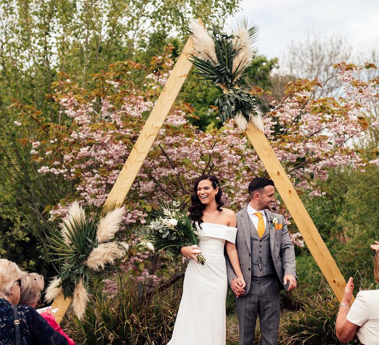 Bride in strapless Vagabond wedding dress and groom in grey Hugo Boss suit stand holding hands under a wooden triangular wedding arch with pampas grass and palm leaf decor