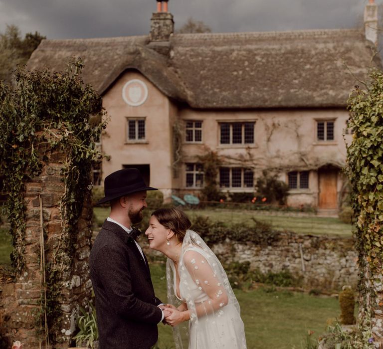 Bride in Charlie Brear wedding dress and daisy chapel length applique veil laughs whilst holding hands with groom in grey tweed suit and black fedora at garden party wedding at Hayne, Devon