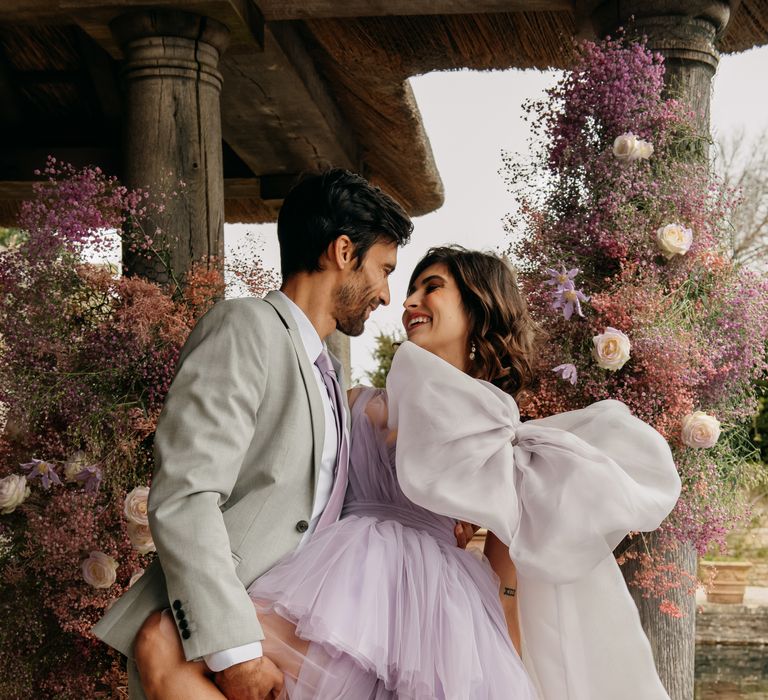 Groom holds bride leg as he leans her back whilst she wears purple chiffon gown