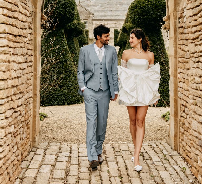 Bride & groom stand under brick archway whilst bride wears short bridal gown with large bow