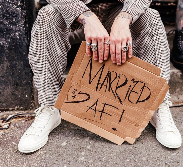 Groom in a check suit and trainers holding a card board sign that says Married AF