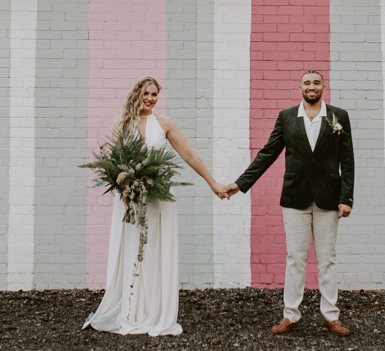 Bride and groom holding hands in front of a pink striped wall, the bride is wearing a wide legged jumpsuit and the groom is wearing a green velvet blazer