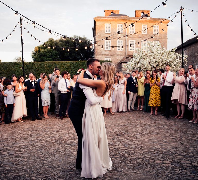 Couple dance outdoors whilst wedding party looks on and lights hang above them 