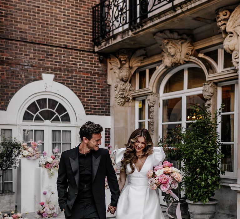 Stylish bride and groom at luxury Mayfair wedding with bride in a Love In Lace Bridal gown with puff sleeves and front split and groom in all black suit and shirt 