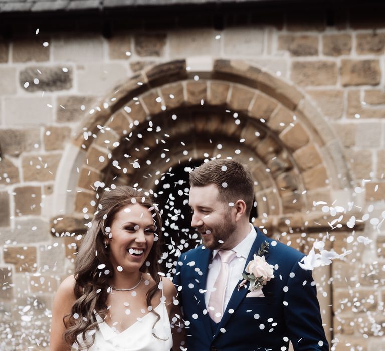 Bride in a white slip wedding dress and groom in a navy suit having confetti thrown over them