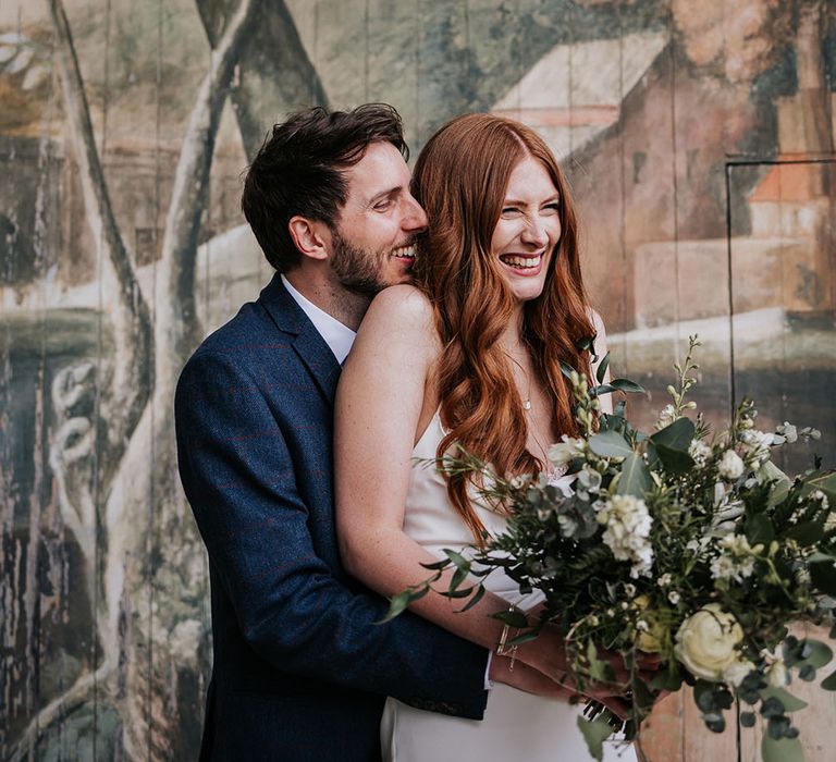 Groom in a navy blue suit embracing his bride in a slip wedding dress on the stage at The Larmer Tree wedding venue's outdoor theatre 