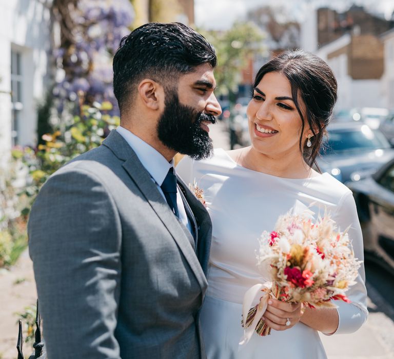 Bride looks lovingly at her groom as they stand in front of house with hanging purple wisteria tree