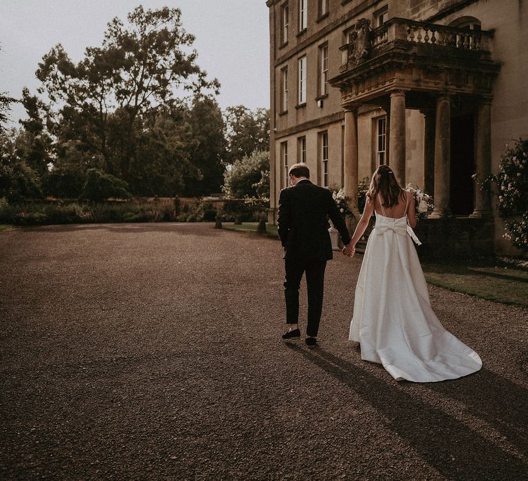 Bride & groom walk hand in hand as bride wears Jesus Peiro dress with bow to the back